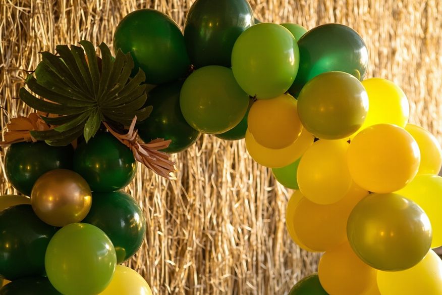 Arco de balões verde e amarelo com folhagens tropicais e painel de fundo de esteira de palha
