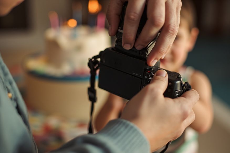 Mãos de fotógrafo segurando câmera na altura de criança, com bolo de aniversário iluminado por velas desfocado ao fundo