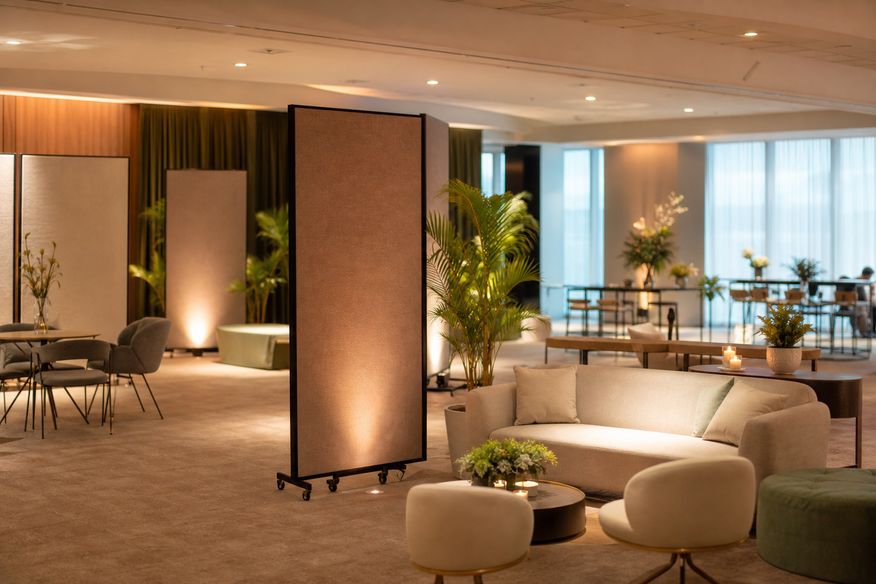 Sala de workshop corporativo com painéis modulares, mobiliário de lounge e plantas.